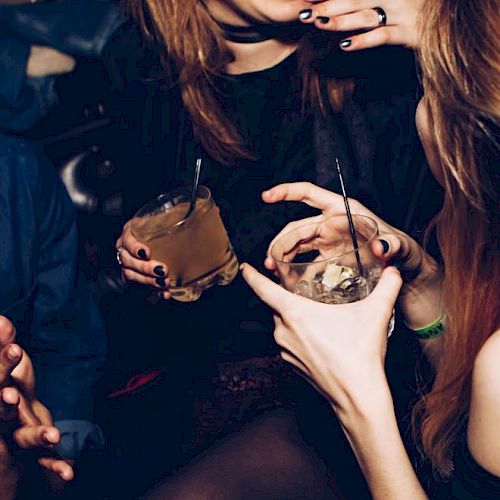 A group of people are socializing, holding drinks, and laughing together in a dimly lit environment, possibly a bar or a party setting.