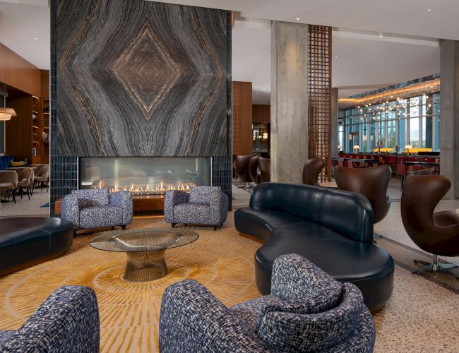 The image shows a modern lounge with a fireplace, stylish seating, marble details, and large windows with a scenic view.