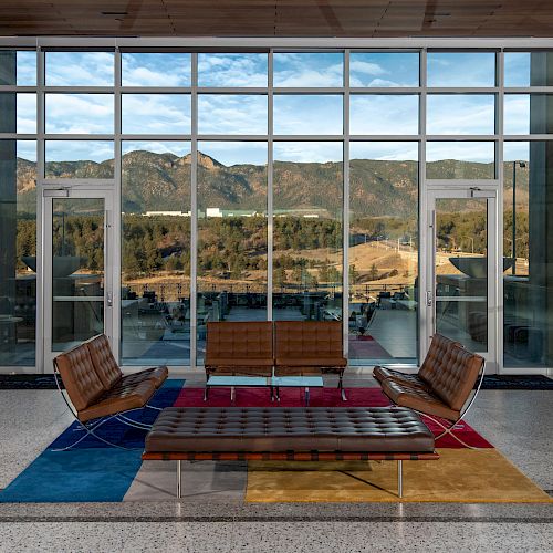 A lounge area with modern brown furniture, large windows showcasing a mountain view, and colorful rugs on the floor.