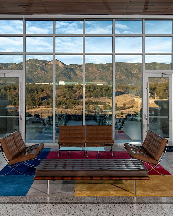 A lounge area with modern brown furniture, large windows showcasing a mountain view, and colorful rugs on the floor.