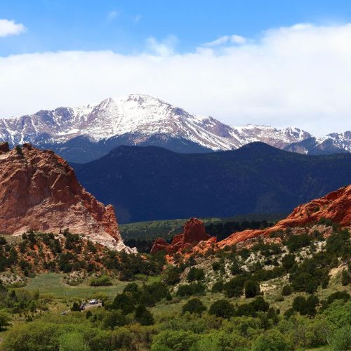 A landscape features red rock formations, lush greenery, and snow-capped mountains under a blue sky with clouds.