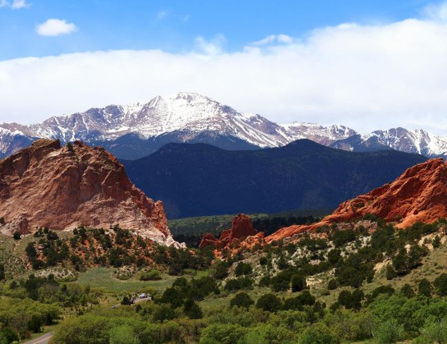 A landscape features red rock formations, lush greenery, and snow-capped mountains under a blue sky with clouds.