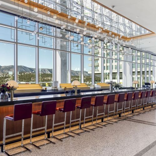 A modern bar with high chairs and large windows showing a scenic view outside. The space features contemporary lighting fixtures.