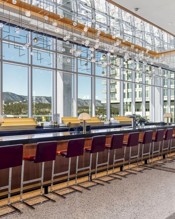 A modern bar with high chairs and large windows showing a scenic view outside. The space features contemporary lighting fixtures.