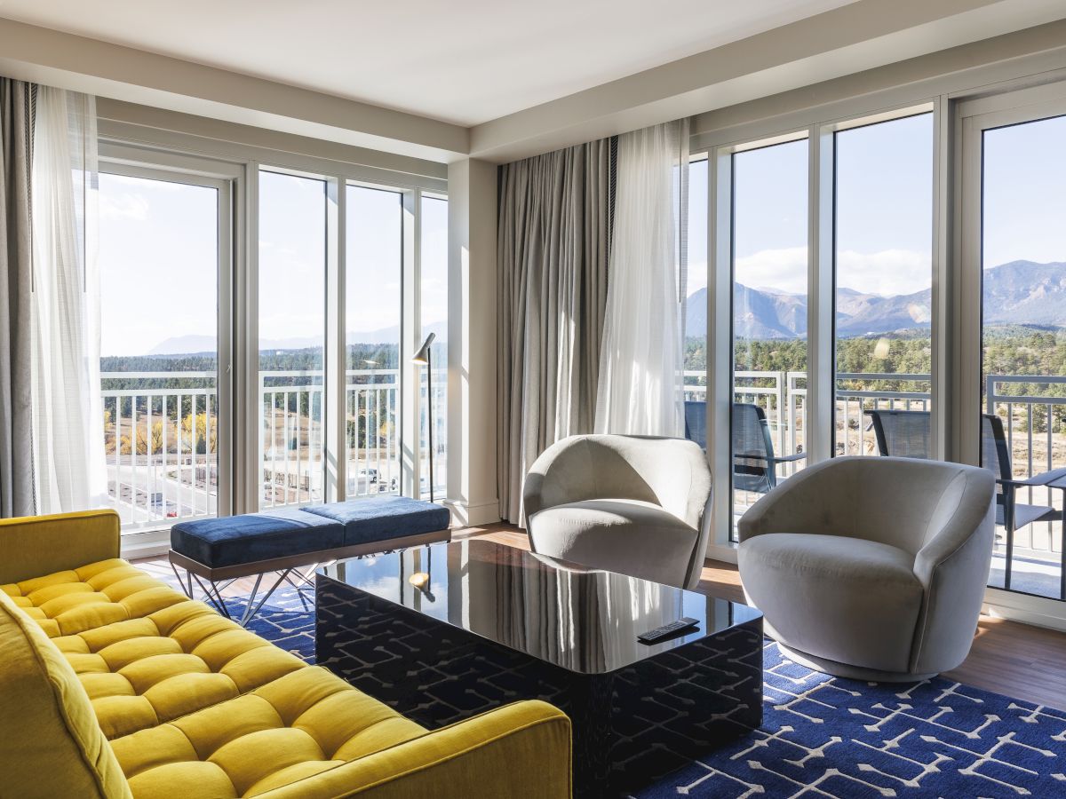 A modern living room with a yellow sofa, two gray chairs, a black coffee table, and large windows with a view of mountains and a balcony.