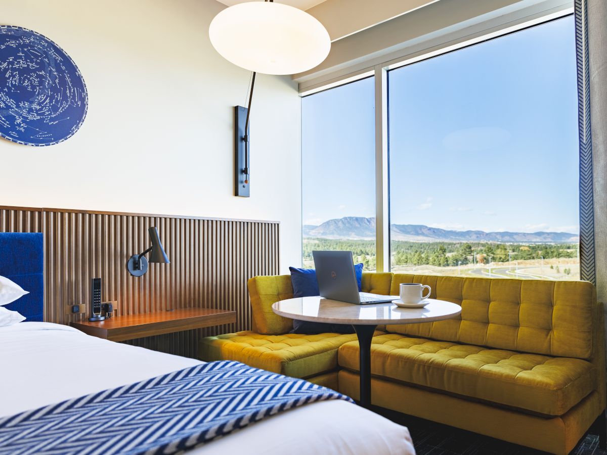 A modern hotel room with a bed, yellow couch, and small round table with a laptop and mug, overlooking a scenic landscape through large windows.