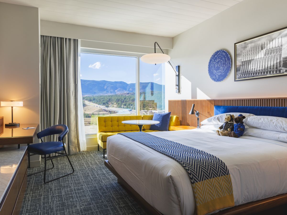 A cozy hotel room with a bed, decorative pillows, a desk, and a window showing a scenic mountain view. Modern decor completes the setting.