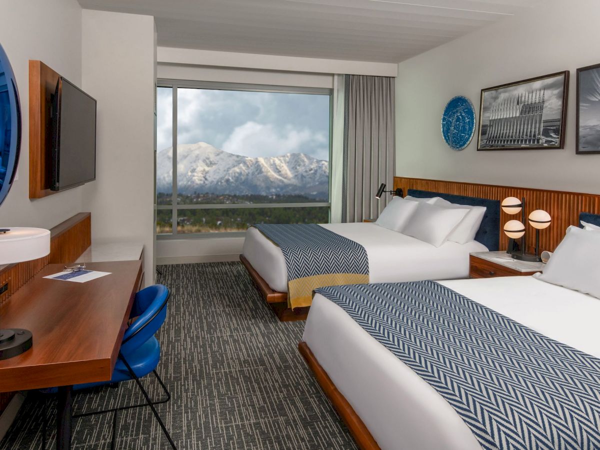 A modern hotel room with two double beds, a desk and chair, a TV, and a large window showcasing a mountain view. Frames and a clock on the wall.