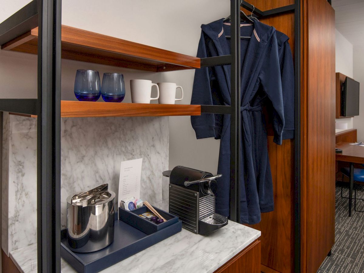 A hotel room refreshment station with a coffee machine, cups, ice bucket, water glasses, a note, and two hanging navy blue robes.