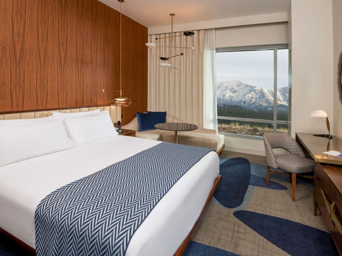 This image shows a modern hotel room with a large bed, a desk, a lounge chair, and a window offering a scenic view of snow-capped mountains.