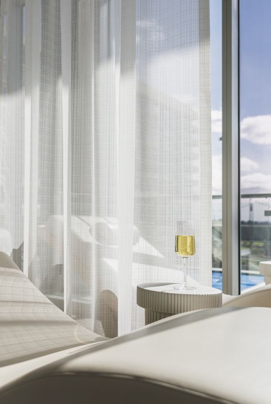 A sunlit room with a modern lounge chair facing large windows, overlooking a balcony with glass railing and a view of the sky and clouds.