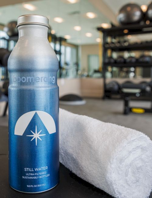 A water bottle and towel are placed on a surface in a gym, with equipment like dumbbells and a treadmill in the background.