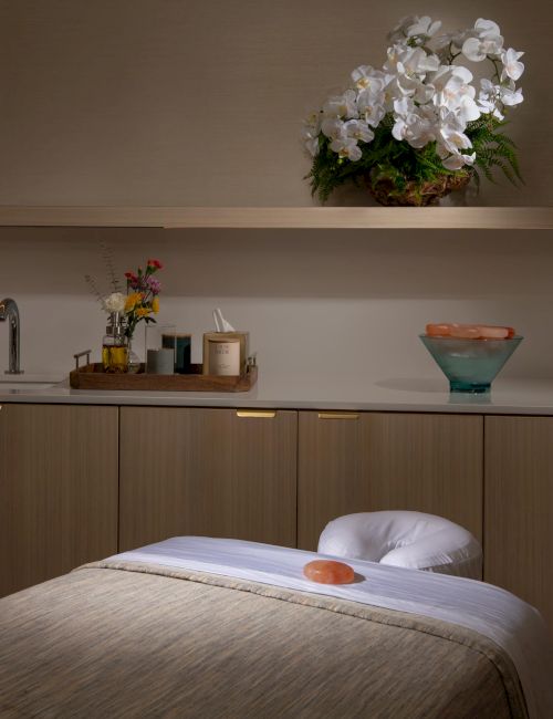 A serene spa room with a massage table, flowers, candles, a bowl of stones, a sink, and elegant orchids on a shelf.