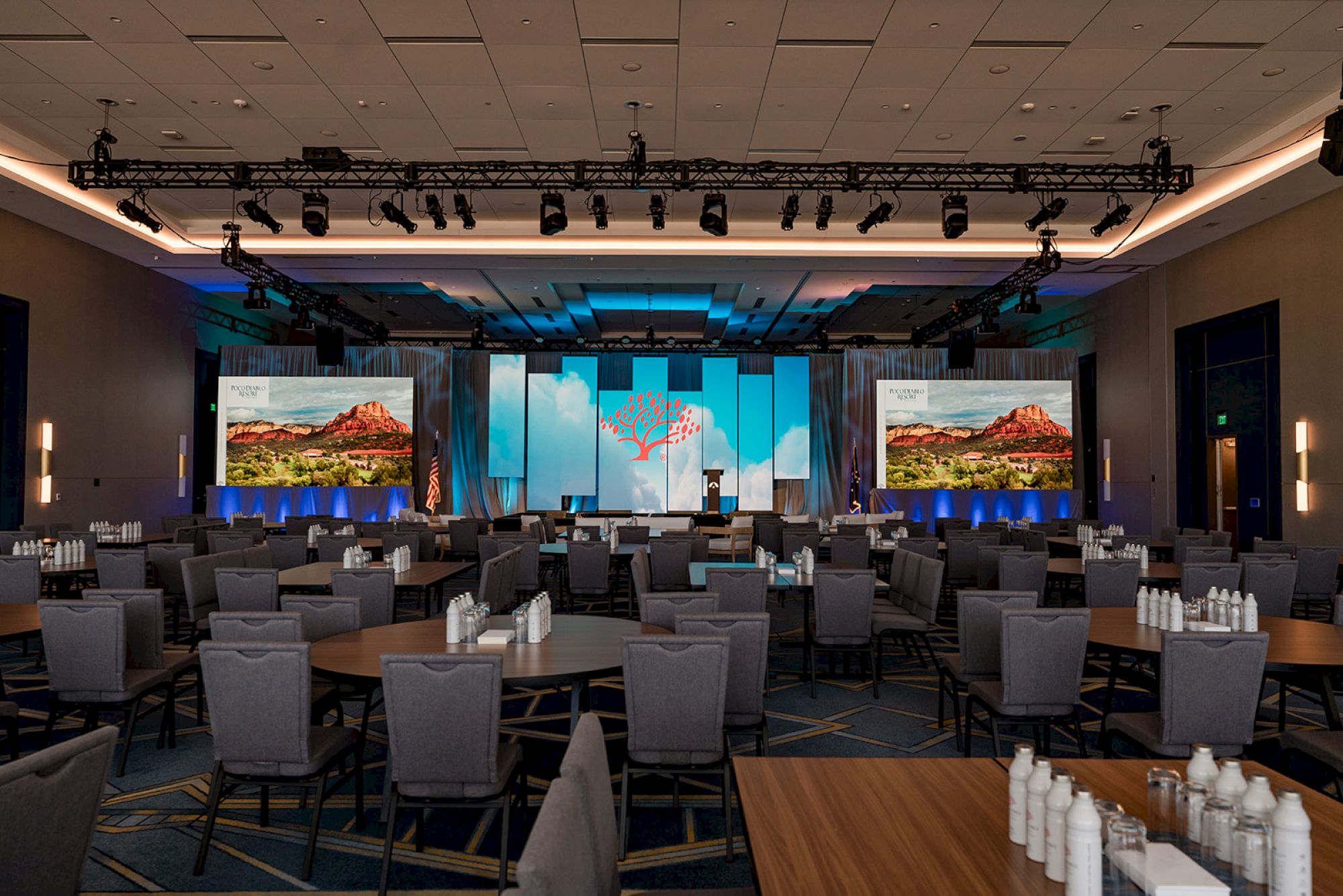 The image shows a conference room setup with rows of tables and chairs, screens displaying scenic landscapes, and bottled water on tables.