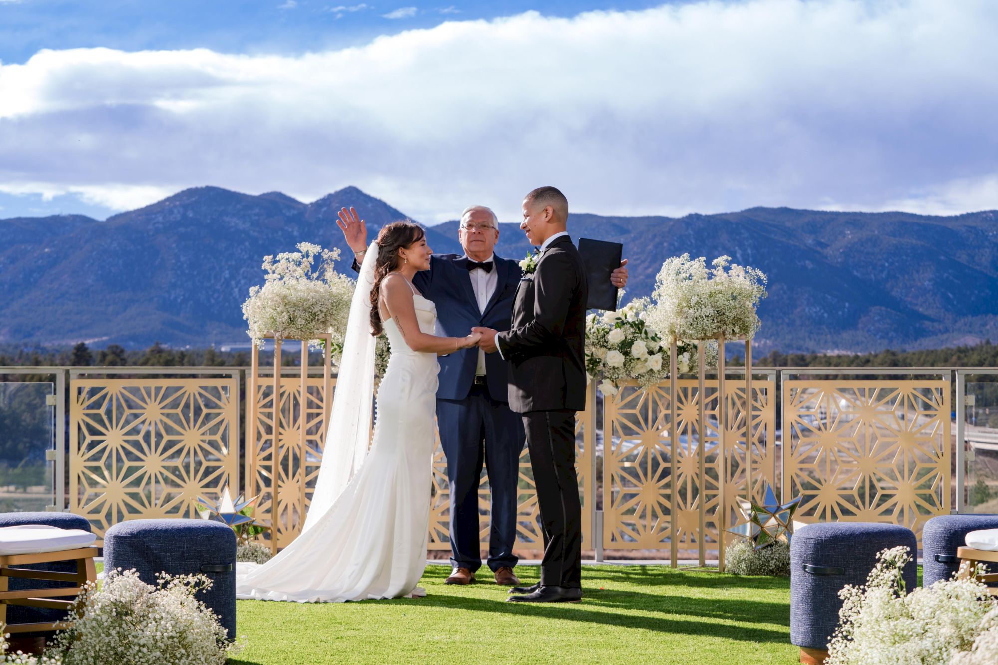 A couple is exchanging vows outdoors with a scenic mountain backdrop. The officiant stands between them on a decorated stage.