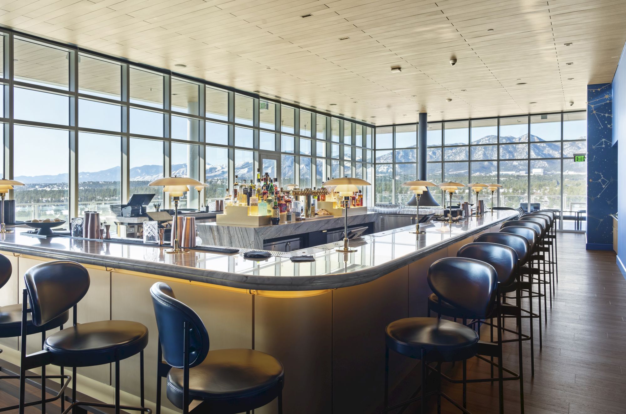 A sleek bar with a marble counter, lined with stools, large windows displaying a mountain view, and bottles arranged behind the bar.