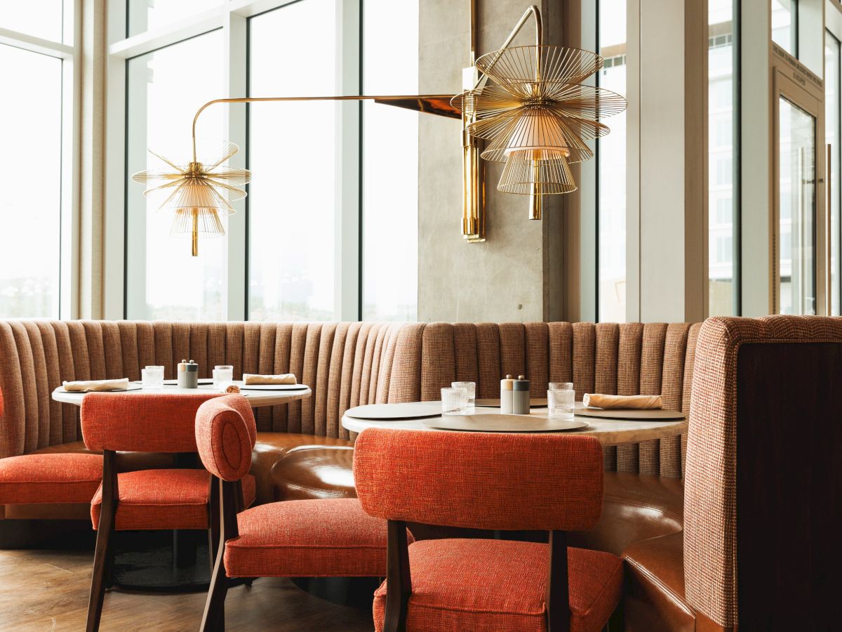 A stylish restaurant interior featuring orange chairs, round tables with place settings, and modern lighting fixtures against tall windows.