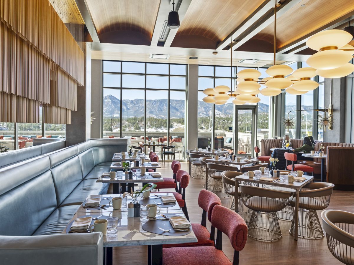 A modern restaurant interior with set tables, red chairs, large windows showing mountain views, and stylish pendant lights hanging from the ceiling.