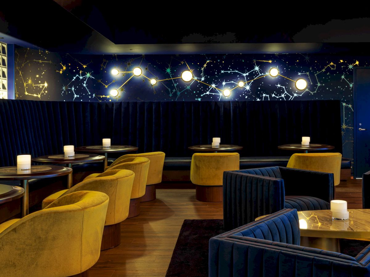 A chic lounge with yellow and blue seating, small tables, and a dark wall displaying a constellation design, softly lit by overhead lights.