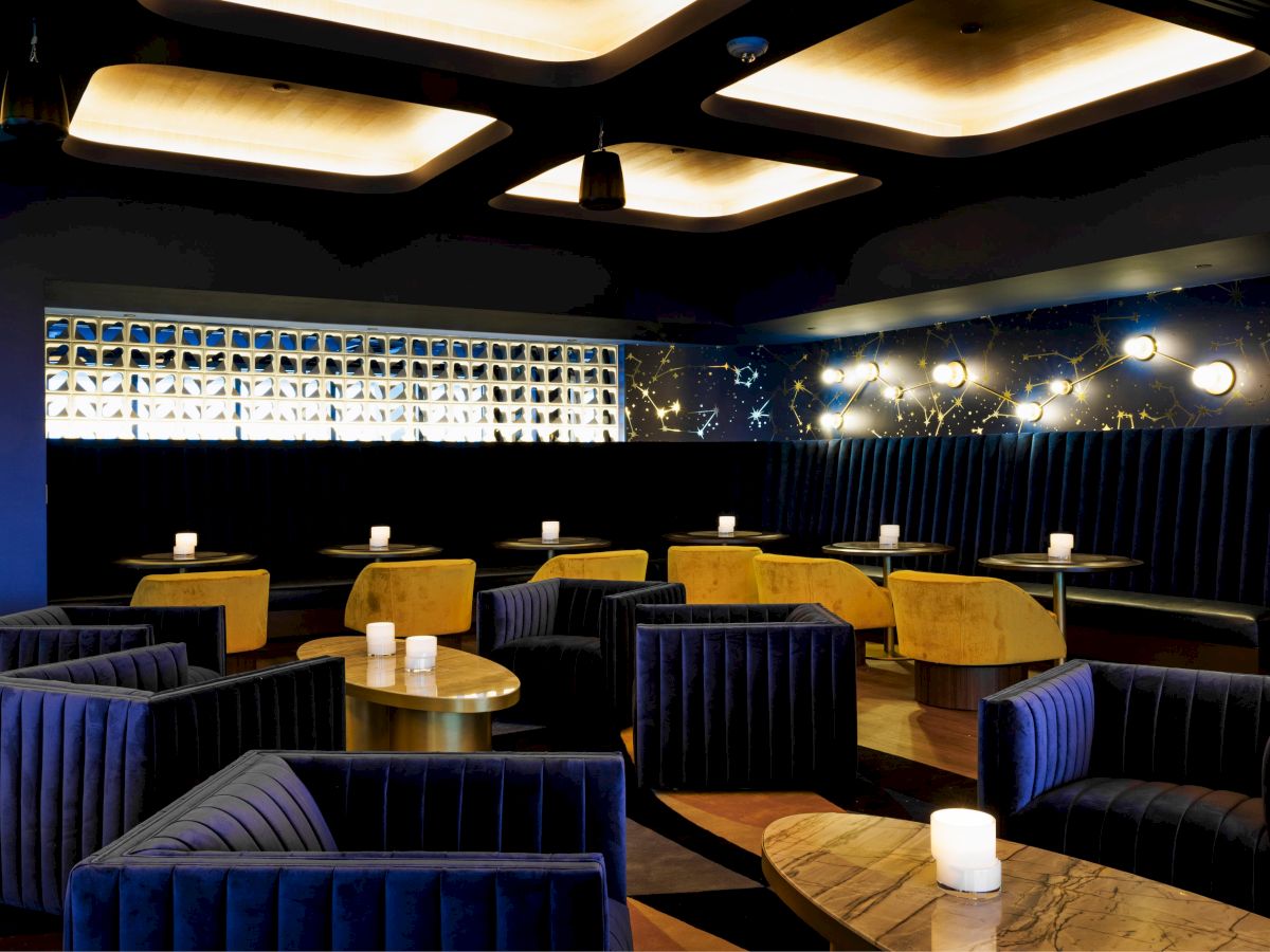 The image shows a stylish lounge area with blue chairs, yellow booths, and marble tables, illuminated by soft lighting on the ceiling.