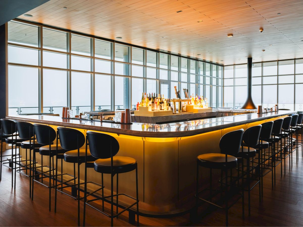 The image shows a stylish bar with a glowing counter, surrounded by high chairs, and large windows offering a bright, open view.