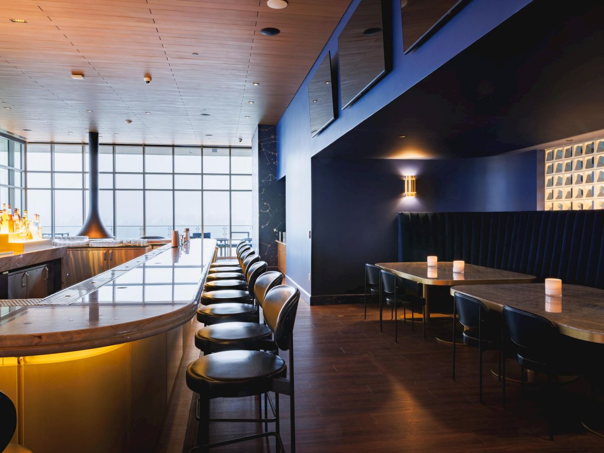 A modern bar interior with stylish lighting, a long counter with stools, and a seating area with candles on tables.