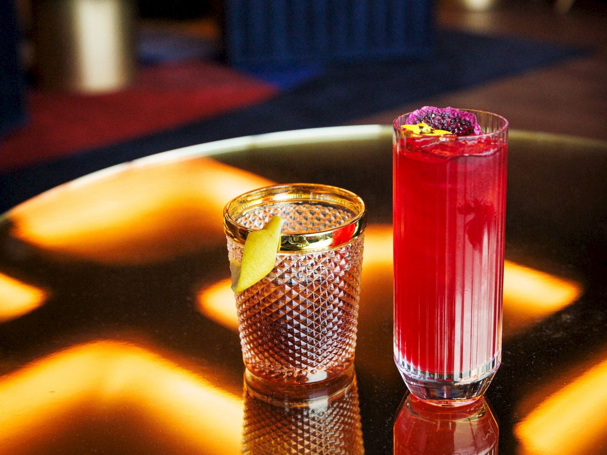 Two drinks on a reflective table: one in a textured glass with a lemon twist, the other a tall red cocktail garnished with a purple flower.