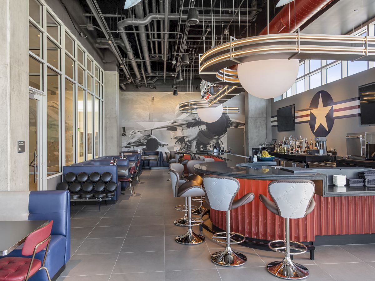 A modern bar with stools, a mural of a jet and star emblem, and seating areas, featuring an industrial ceiling and contemporary lighting fixtures.