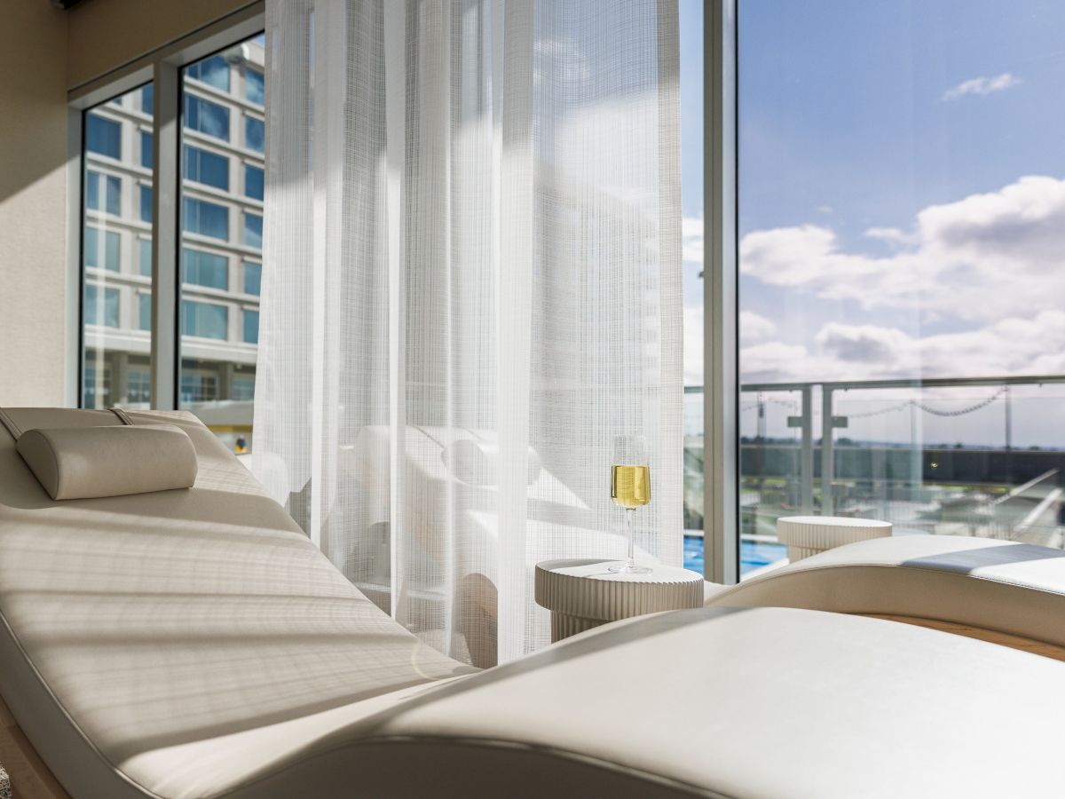 A sunny room with a lounge chair, sheer curtains, and a view of the sky and neighboring building through large windows.