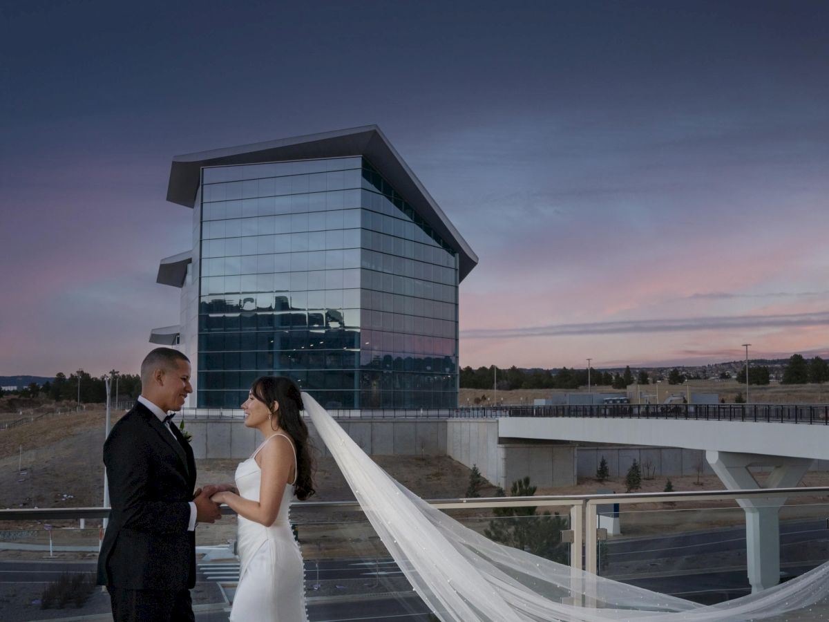 A couple in formal attire stands on a terrace at dusk, with a modern building in the background and a long flowing veil.