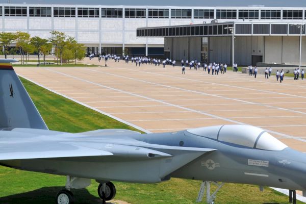 The image shows a military aircraft displayed on a grassy area in front of a large building, with people walking in the background.