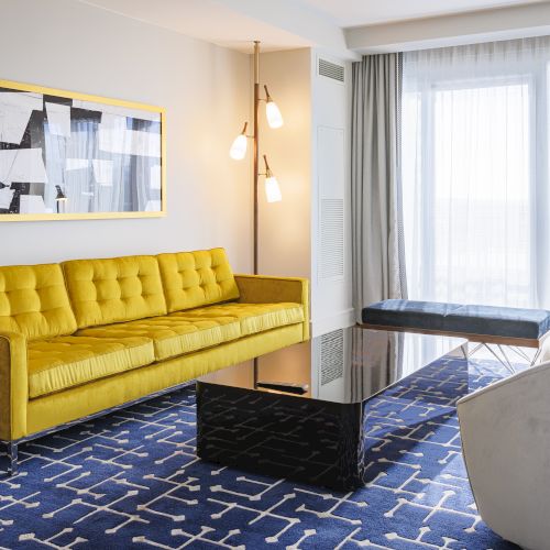 A modern living room with a yellow sofa, abstract art, a black coffee table, blue patterned rug, and large windows with sheer curtains.