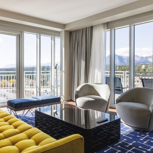 A modern living room with a yellow sofa, two round chairs, and a black coffee table, overlooking a balcony with mountain views.