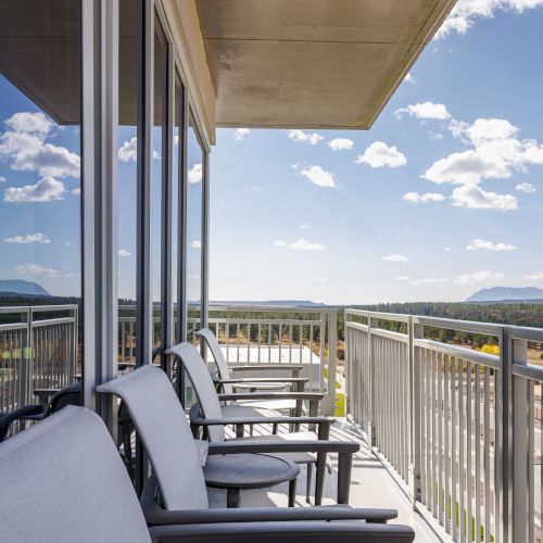 A balcony with chairs overlooks a scenic landscape, featuring clear skies and distant mountains, with reflective glass windows beside it.