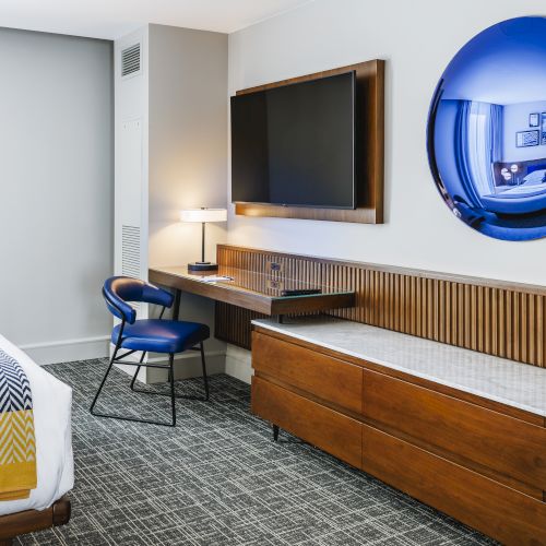 A modern hotel room featuring a bed, chair, desk, flat-screen TV, lamp, and a round wall mirror with a blue tint.