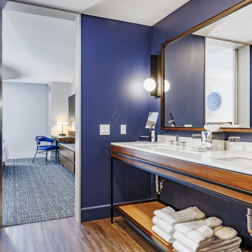 The image shows a modern bathroom with a double sink, wooden accents, and a mirror, leading into a room with a chair and desk.