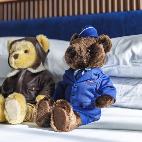 Two teddy bears dressed as pilots sit on a bed with white pillows, featuring brown and blue outfits.