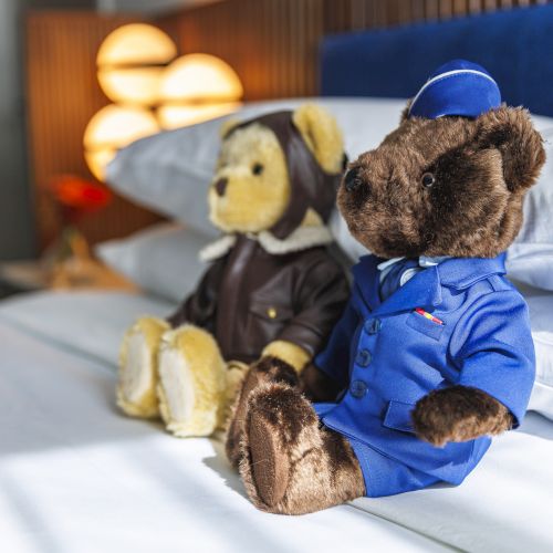 Two teddy bears dressed as pilots sit on a neatly made bed, with a stylish lamp and headboard in the background.