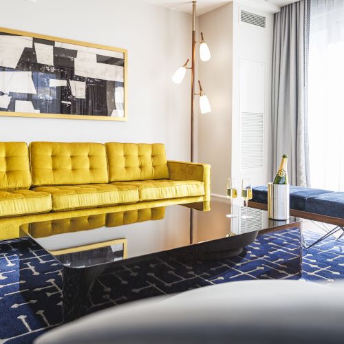 A modern living room with a yellow sofa, abstract art, and a black coffee table. Decor includes a blue patterned rug and champagne bottle.