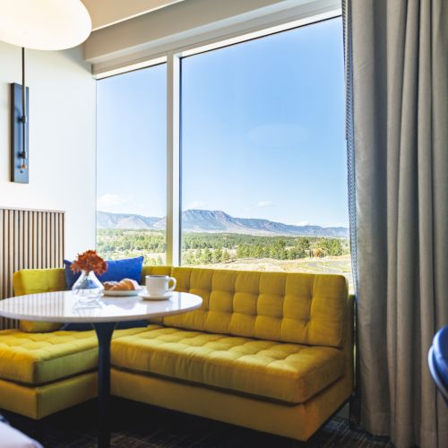 A cozy hotel room with a yellow sofa by a large window, offering a scenic mountain view. A round table with a cup and vase is nearby.