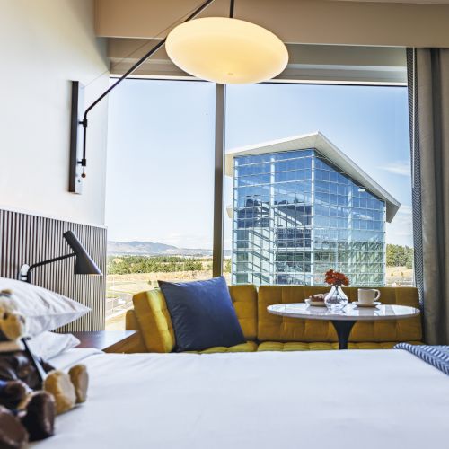 A cozy hotel room with teddy bears on the bed, a seating area, and a city view through a large window.