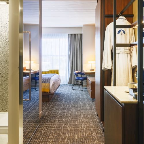 This image shows a hotel room interior with a bed, desk, open wardrobe with robe, and a small coffee station, viewed from the entrance.