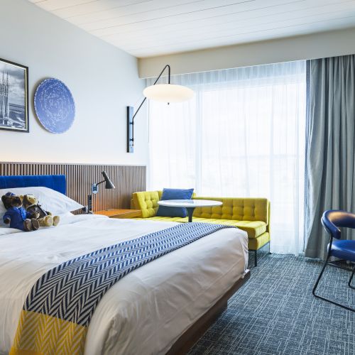 A modern hotel room with a bed, desk, chair, and a small seating area by the window. Bright, cozy, and neatly arranged.