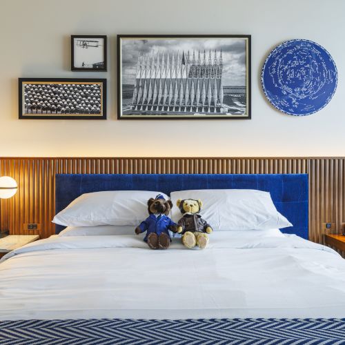 A neatly made bed with two stuffed bears, surrounding nightstands with lamps, and framed art on the wall above the headboard.