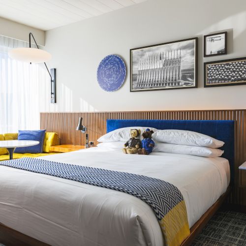 A neatly decorated hotel room featuring a bed with blue and yellow accents, wall art, a cozy seating area, and two teddy bears on the bed.