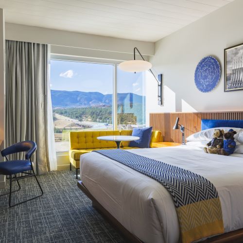 A cozy hotel room features a bed, a desk, a chair, a sofa, art on the walls, and a window with a scenic mountain view, creating a relaxing space.