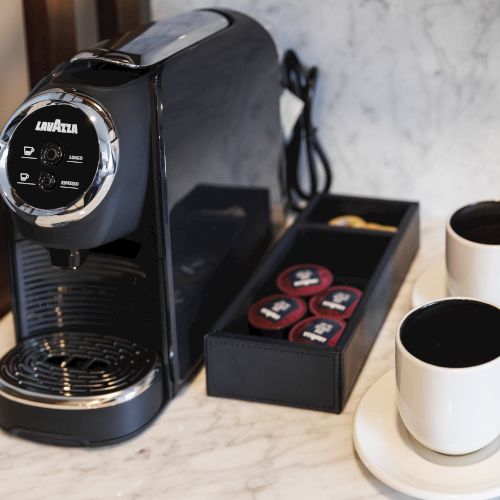 A black coffee machine with pods beside it, set on a marble surface. Two white cups on saucers are filled with black coffee.