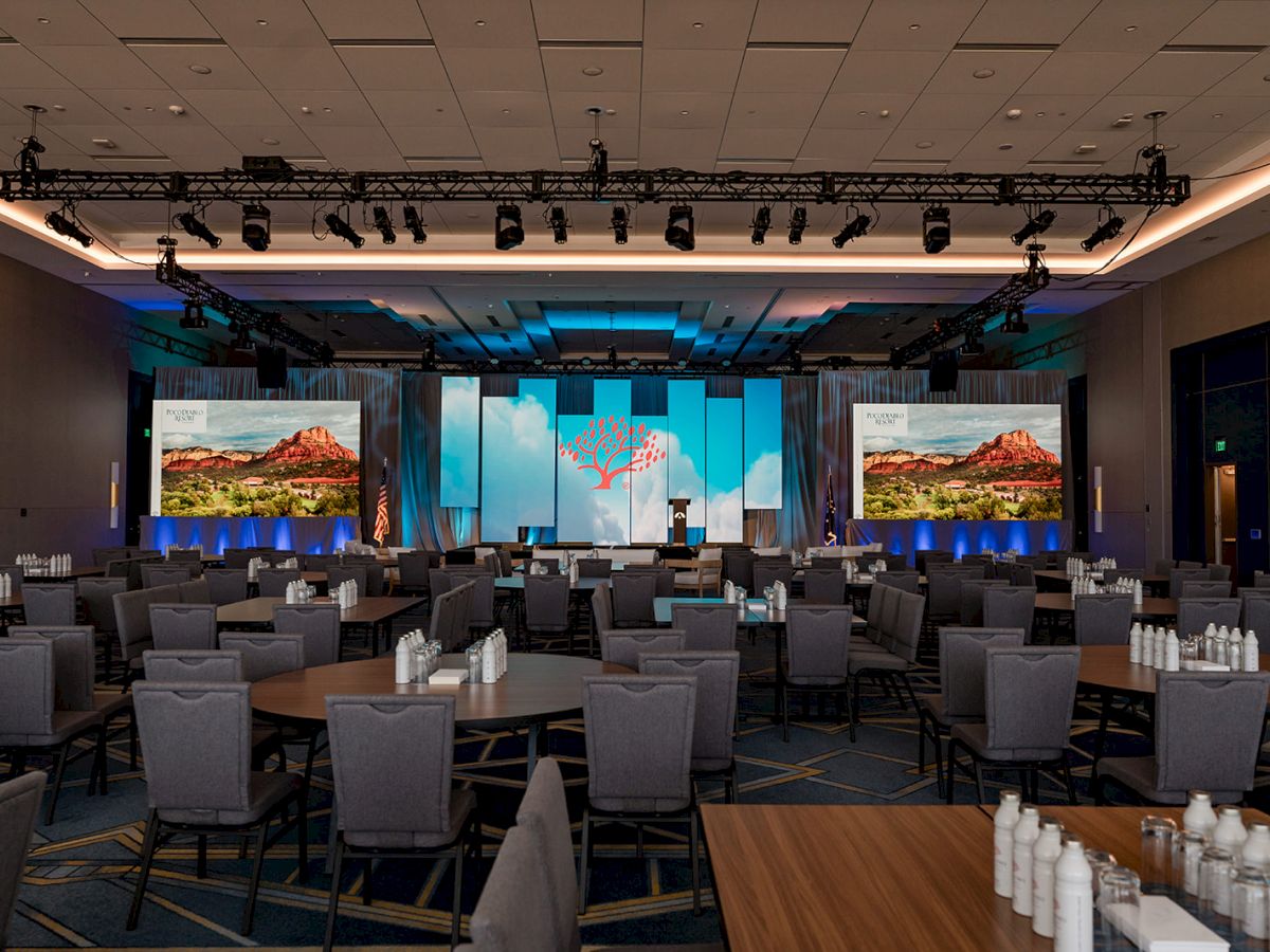 A conference room with round tables, chairs, water bottles, and a stage featuring screens and lighting setup displayed in the image.