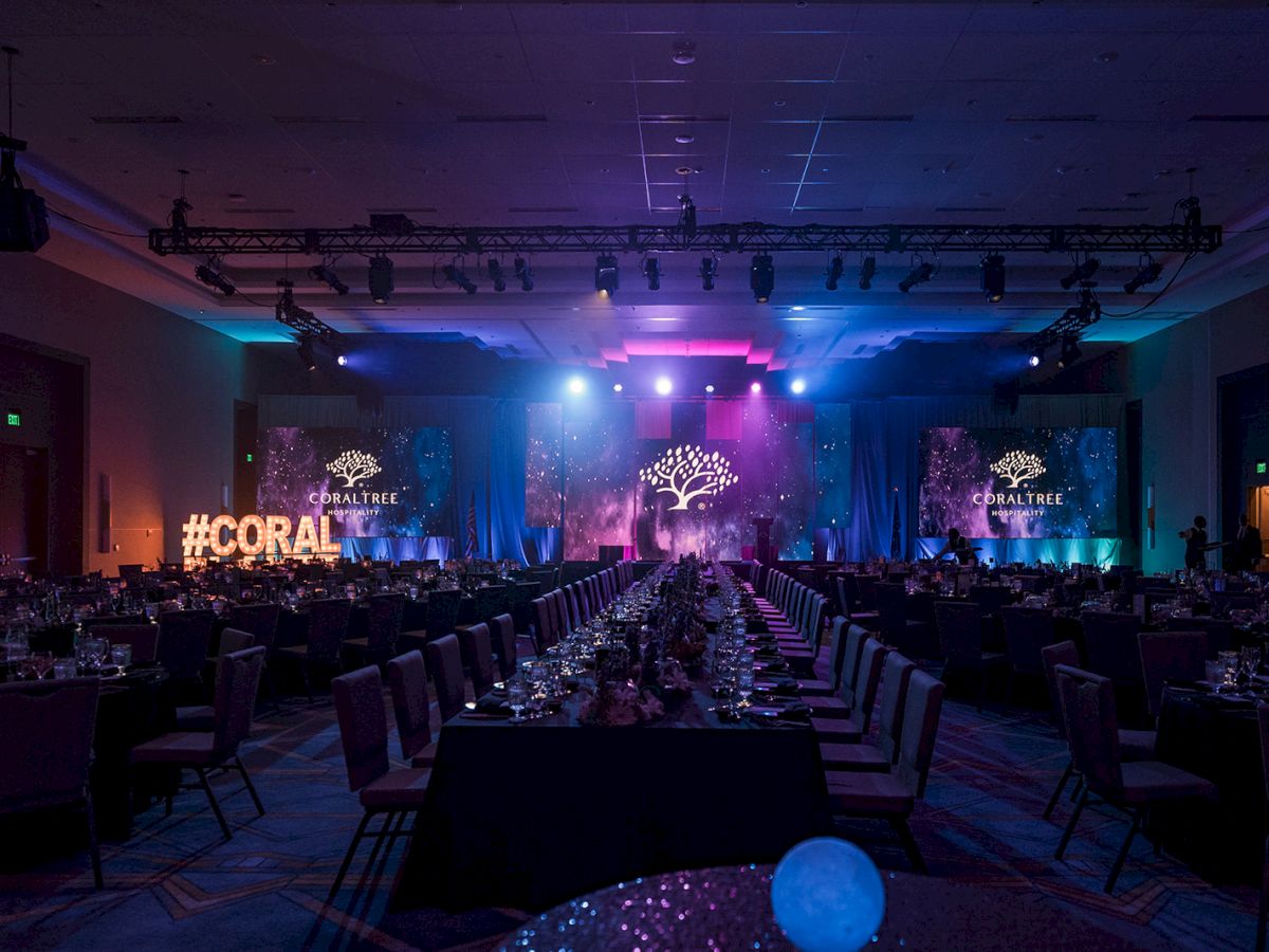 A banquet hall with elegant lighting, set for an event. The room has a "#CORAL" sign and "CoralTree Hospitality" logos on screens.
