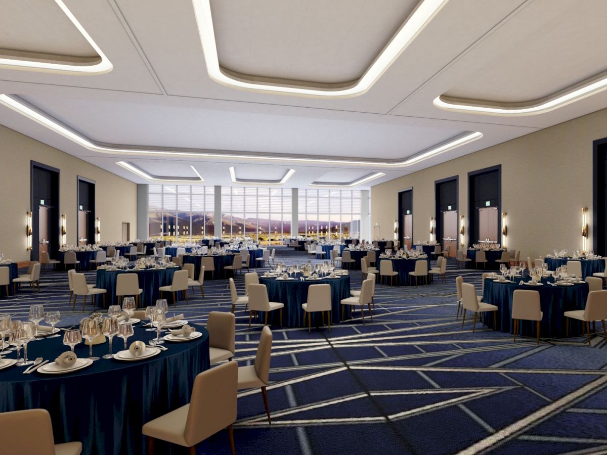 A large banquet hall with round tables set for an event, featuring modern lighting, carpeted flooring, elegant place settings, and large windows at the end.